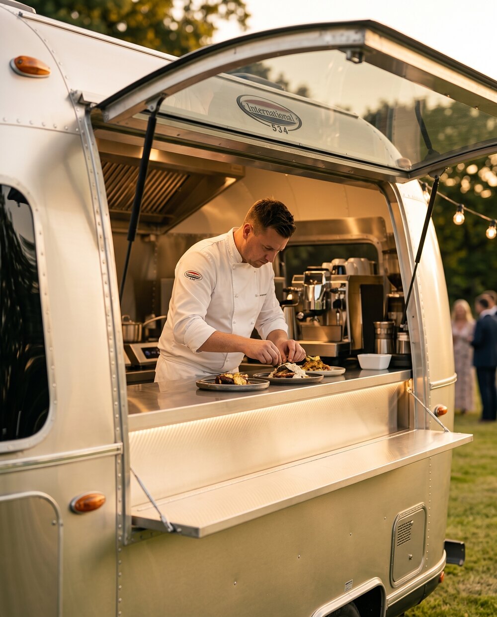 Blick in die geöffnete Ausgabeklappe des Airstream: Ein Koch richtet ein Gericht auf der Edelstahltheke an.